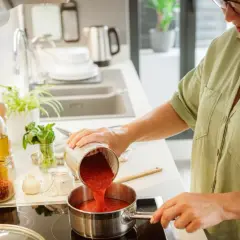Mujer vertiendo tomate triturado en una olla para espesar una salsa. Ingredientes como aceite, ajo y especias a la vista.