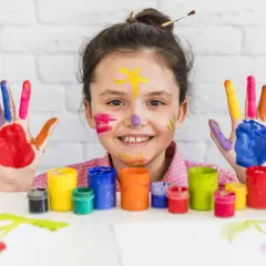 Uśmiechnięta dziewczynka z kolorowymi dłońmi i twarzą. To przykład, jak arteterapia co to znaczy – radość tworzenia.