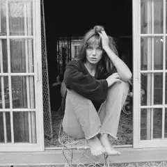 Jane Birkin mit typischen 70er Frisuren, lässig auf einem Stuhl sitzend, vor einer Tür mit Sprossenfenstern.