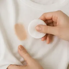 Mano aplicando disco de algodón sobre mancha de maquillaje en camiseta blanca. Aprende como quitar manchas de maquillaje de la ropa.