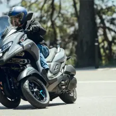 Hombre en casco azul conduce un triciclo Yamaha Tricity 500 por una carretera arbolada, inclinándose en una curva.