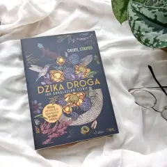 Okładka książki "Dzika droga" z motywami roślinnymi i zwierzęcymi, obok okulary i inne książki.