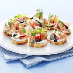 Köstliche italienische Crostini mit Prosciutto, Feigen, Mozzarella und Rucola auf einem weißen Teller.