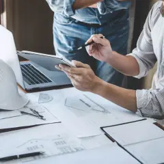 Architekci analizują plany, przygotowując się do uprawomocnienia pozwolenia na budowę. Na stole leżą kask, laptop i dokumenty.