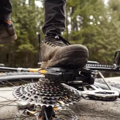 A mountain biker's muddy shoe rests on the SRAM T-Type derailleur and cassette of a bike lying on its side.