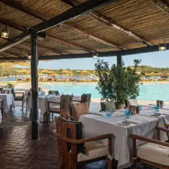 Tavoli apparecchiati sotto un tetto di paglia, con vista sulla piscina e sulla spiaggia del **hotel Cala di Volpe**.