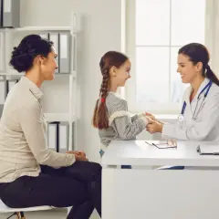 Uśmiechnięta lekarka, hematolog, trzyma za ręce małą pacjentkę, podczas gdy jej mama siedzi obok.