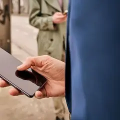 Dłoń trzyma smartfon z ciemnym ekranem. W tle druga osoba również korzysta z telefonu.