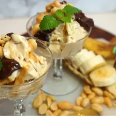 Två glas med nyttig glass, toppad med choklad, jordnötter och mynta. Bananskivor och jordnötter ligger bredvid.