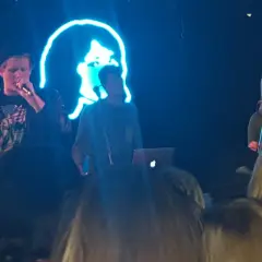 Koncert zespołu tv girl. Na scenie trójka artystów, w tle neonowa podobizna twarzy.