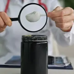Persona con bata blanca examina con lupa polvo blanco de un envase, investigando creatina efectos secundarios.
