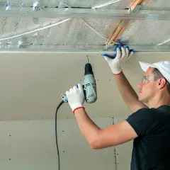 Un ouvrier installe des isolants minces sous un faux plafond en plaques de plâtre, utilisant une perceuse pour fixer les éléments.