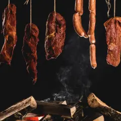 Wędzone mięsa i kiełbasy wiszą nad ogniskiem, tworząc idealne warunki do wędzenia. Dym unosi się, nadając potrawom niepowtarzalny aromat.