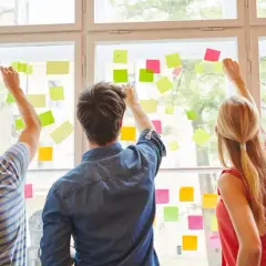 Fünf Personen planen an einem Fenster mit bunten Haftnotizen. Sie arbeiten an ihrer kaufmännischen Ausbildung nebenberuflich.