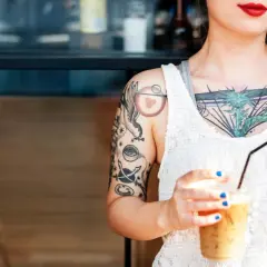 Junge Frau mit vielen Tattoos, darunter ein Vogel und ein Auge. Sie hält einen Eiskaffee. Ein Tattoo ab 16, das ihre Persönlichkeit unterstreicht.