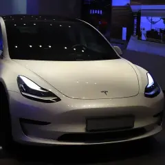 Biały samochód Tesla Model 3 z włączonymi światłami, prezentowany w salonie.