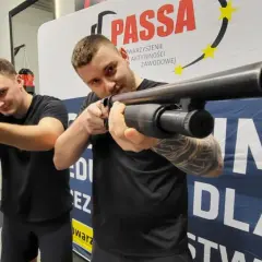 Dwóch mężczyzn ćwiczy strzelanie z broni gładkolufowej. Jeden z nich, kwalifikowany pracownik ochrony, celuje w obiektyw.