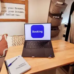 Laptop con logo Booking aperto su un tavolo di legno, accanto a un quaderno "Happy Thoughts". Pianifica il tuo prossimo viaggio con credito booking.