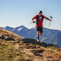 Ein Mann im roten Shirt und schwarzer Hose läuft mit Stöcken einen Bergpfad entlang. Sein intensives anaerobes Training fordert ihn in der atemberaubenden Berglandschaft.
