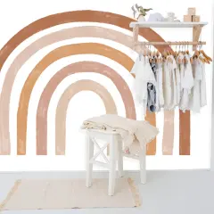 Un arc-en-ciel aux tons terreux est peint sur un mur blanc, orné d'une penderie d'enfants et d'une décoration murale en macramé.