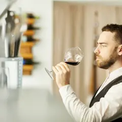 Mężczyzna w białej koszuli i kamizelce, sommelier, kto to? Wącha kieliszek czerwonego wina, oceniając jego aromat.