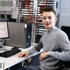 Młody mężczyzna w szarym swetrze siedzi przy biurku z komputerem w sali lekcyjnej.