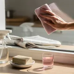 Mano che pulisce un vetro con un panno rosa, mostrando come pulire i vetri senza aloni con sapone di Marsiglia e spray.
