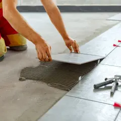 Un operaio in ginocchio posa una piastrella su cemento fresco, creando un pavimento con posa piastrelle a 45 gradi. Si vedono livellatori rossi e uno strumento per piastrelle.