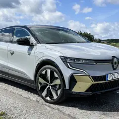 En vit Renault Megane E-Tech står parkerad på en grusväg med en gul och grön fält i bakgrunden. En del av en testkörning.