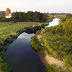 Malownicza wieś nad Liwcem, z charakterystycznym dworem i wieżą, otoczona zielonymi łąkami i drzewami. Rzeka Liwiec meandruje przez krajobraz.