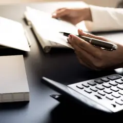 Ręce trzymają długopis nad dokumentami, obok kalkulator i laptop. Analiza danych, by dowiedzieć się, ile zarabia dietetyk.