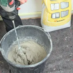 Personen använder en borrmaskin för att blanda betong i en hink. En gul säck med Skalflex-märkning står bredvid.