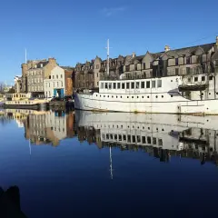 Barcos y edificios históricos reflejados en el agua en Leith, Edimburgo. El "Ristorante De Niro" en un barco blanco destaca.