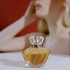 Elegancka buteleczka perfum z płynem o barwie **citral** na białym tle, w tle niewyraźna sylwetka kobiety.