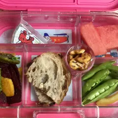 A pink bento box with watermelon, peas, pretzels, bread, and veggie sticks. Add an ice pack to keep this bento box cold.