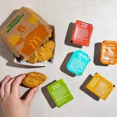 Ręka sięga po jednego z chrupiących McNuggetsów z pudełka. Obok leży zestaw sosów: Tangy BBQ, Spicy Buffalo, Ranch, Sweet 'N Sour i Honey Mustard.