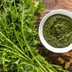 Un mazzo di **tarassaco foglie** fresche accanto a una ciotola di pesto verde brillante e pasta fusilli su un tavolo di legno rustico.