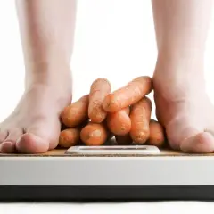 Stopy na wadze, a między nimi stos marchewek. Waga stoi w miejscu, symbolizując dietę i zdrowe nawyki.