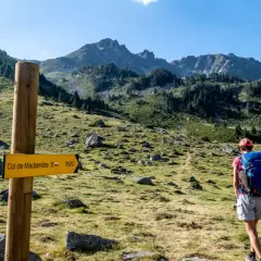 Randonneuse sur le GR10, suivant le panneau jaune indiquant le Col de Madamète. Paysage de montagne verdoyant.