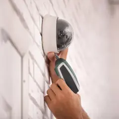 Installation d'un capteur infra rouge de surveillance, une main utilise un tournevis électrique pour fixer la caméra dôme sur un mur en briques blanches.
