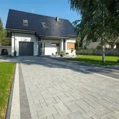 Nowoczesny podjazd przed domem z szarej kostki brukowej, otoczony zielonym trawnikiem i drzewami.