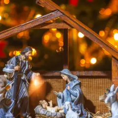 Szopka bożonarodzeniowa z figurkami Świętej Rodziny, pasterzy i zwierząt. W tle migoczą światełka choinkowe, tworząc ciepłą atmosferę.