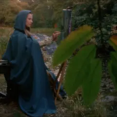 Essy Persson, klädd i en blå kappa, målar vid en sjö.