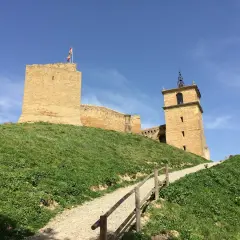 El castillo de San Vicente de la Barquera se alza majestuoso sobre una colina verde, con su torre y murallas de piedra bajo un cielo azul.