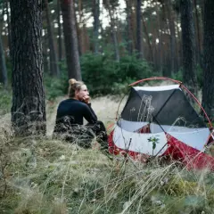 Ein Mann sitzt nachdenklich neben einem roten Zelt im Wald. Hier darf man im Wald zelten und die Natur genießen.