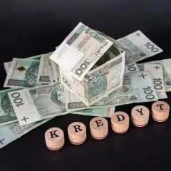 Dom z banknotów 100 zł, symbolizujący kredyt hipoteczny. Słowo "KREDYT" ułożone z drewnianych pieczątek.