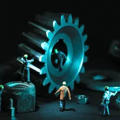Dział utrzymania ruchu pracuje nad wielką zębatką. Małe figurki robotników w kaskach porządkują śruby i nakrętki.