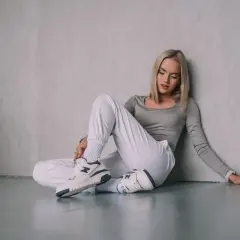Blondynka w białych spodniach i szarym topie siedzi na podłodze, opierając się o ścianę. Na nogach ma stylowe snekarsy.