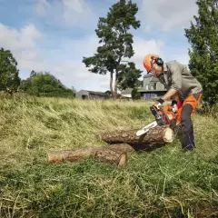 Mężczyzna w kasku i słuchawkach tnie drewno piłą łańcuchową Husqvarna 450.