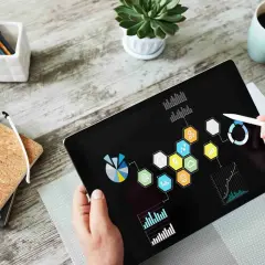 Ręka z rysikiem wskazuje na tablet z wykresami i ikonami, prezentując przykładowe mierniki realizacji celów.
